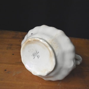 May include: A white ceramic creamer with a ruffled edge, viewed from the bottom. The creamer has a small handle and a slightly stained base. The bottom of the creamer has a maker's mark.
