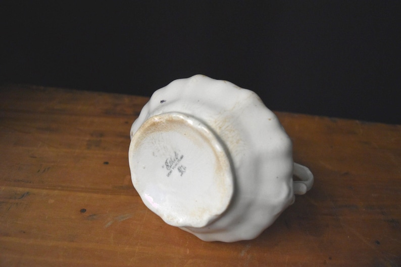 May include: A white ceramic creamer with a ruffled edge, viewed from the bottom. The creamer has a small handle and a slightly stained base. The bottom of the creamer has a maker's mark.