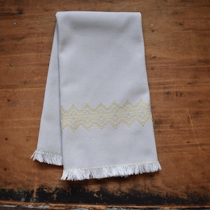 May include: A light gray tea towel with a yellow and white geometric pattern. The towel has a fringed edge.