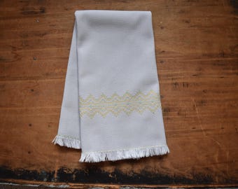May include: A light grey tea towel with a yellow and white geometric pattern. The towel has a fringed edge.