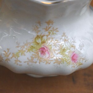 May include: A white porcelain creamer with a scalloped edge and two handles. The creamer is decorated with pink roses and gold floral accents. The creamer is a vintage piece.
