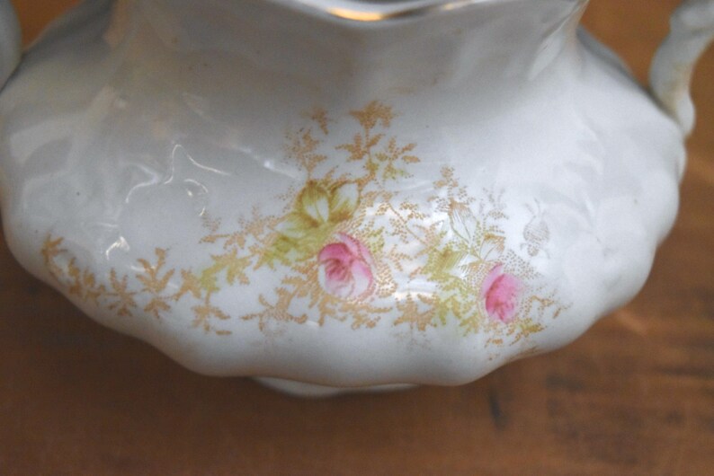 May include: A white porcelain creamer with a scalloped edge and two handles. The creamer is decorated with pink roses and gold floral accents. The creamer is a vintage piece.