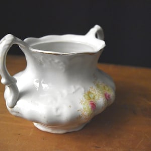 May include: White ceramic creamer with two ornate handles and a scalloped rim. The creamer features a floral design in yellow and pink. The creamer is placed on a wooden surface.