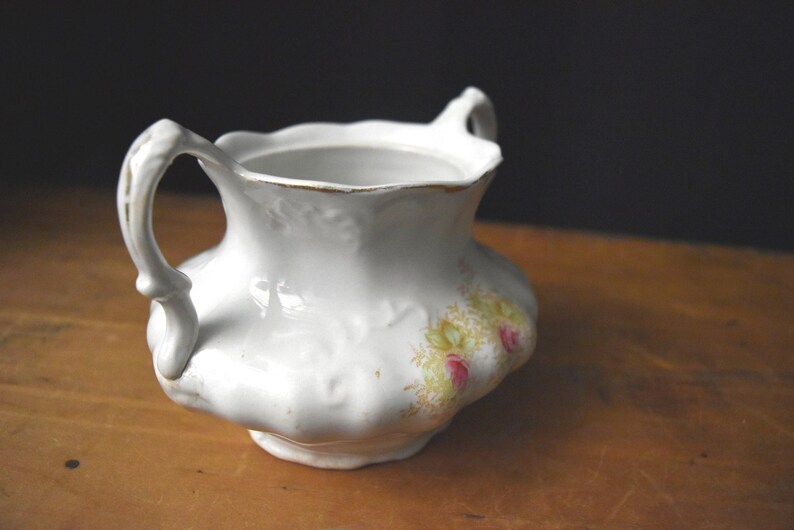 May include: White ceramic creamer with two ornate handles and a scalloped rim. The creamer features a floral design in yellow and pink. The creamer is placed on a wooden surface.