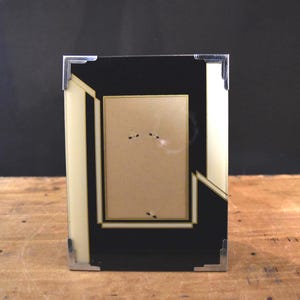 May include: A rectangular picture frame featuring a black and cream geometric pattern. The frame has metallic silver-toned corner accents and a central rectangular opening for a photograph, surrounded by a thin gold-colored border detail.