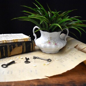 May include: A still life arrangement featuring a white floral vase with green foliage, a stack of old books, and three antique keys. The scene includes rolled sheet music and small, scattered objects.