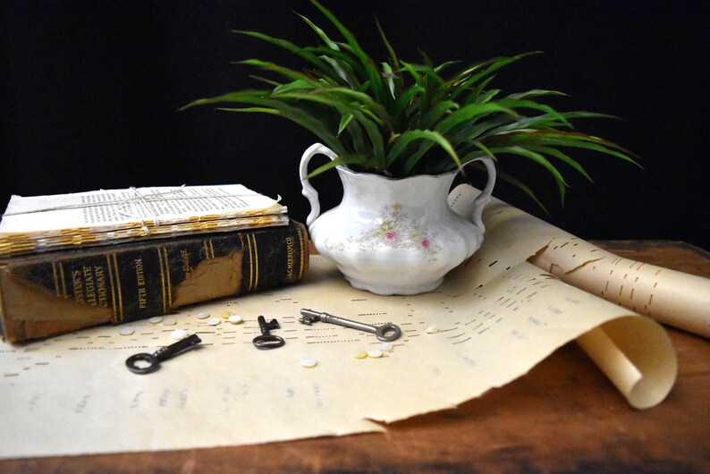 May include: A still life arrangement featuring a white floral vase with green foliage, a stack of old books, and three antique keys. The scene includes rolled sheet music and small, scattered objects.