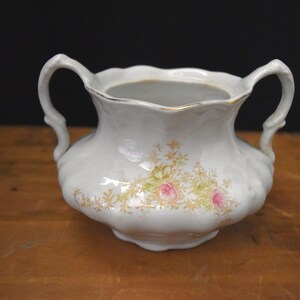 May include: White ceramic sugar bowl with two handles and a scalloped rim. The bowl is decorated with a floral pattern featuring pink roses and gold accents. The sugar bowl sits on a wooden surface.