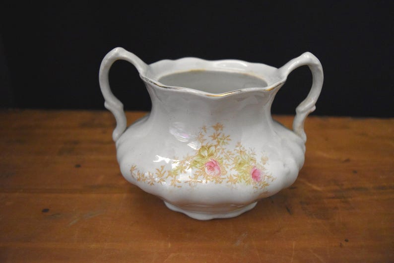 May include: White ceramic sugar bowl with two handles and a scalloped rim. The bowl is decorated with a floral pattern featuring pink roses and gold accents. The sugar bowl sits on a wooden surface.