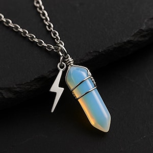 May include: A silver necklace with a light blue and yellow crystal pendant, wire-wrapped, and a lightning bolt charm. The chain and charms are set against a dark background, accentuating the pendant's colour and shape.