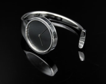 Peut inclure: Une montre bracelet argentée avec un cadran noir et des aiguilles argentées. La montre est sur un fond noir.