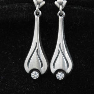 May include: A pair of silver-toned drop earrings featuring a teardrop-shaped metal body with a brushed finish and engraved curved lines. Each earring is topped with a small floral-shaped connector and finished with a single clear round gemstone.
