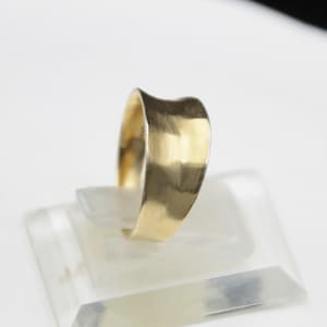 May include: A gold ring with a wide, smooth band featuring a subtle, concave wave-like indentation along the top edge. The metal surface has a soft, brushed finish that reflects light evenly across its curved, continuous form.