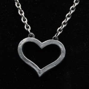 May include: A silver-toned metal necklace featuring an open heart-shaped pendant suspended from a thick, textured link chain. The pendant has a matte, slightly distressed finish with visible surface markings and a simple, hollow center design.