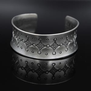 May include: A wide silver-toned metal cuff bracelet featuring a brushed finish and a repeating embossed pattern of small circles and diamond shapes with cross-hatch detailing centred along the band. The bracelet has an open back design.