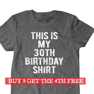 Puede incluir: Camiseta gris con texto blanco que dice "This is my 30th Birthday Shirt". La parte inferior de la camiseta tiene una pancarta roja que dice "Buy 3 Get the 4th Free".