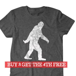 Puede incluir: Una camiseta gris oscuro jaspeada con una silueta blanca de Bigfoot caminando. El texto "BUY 3 GET THE 4TH FREE" est&aacute; impreso en rojo en la parte inferior de la camiseta.
