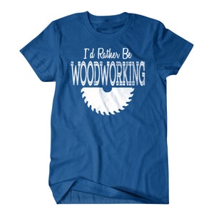 以下が含まれることがあります： ロイヤルブルーのTシャツに、白いグラフィックで「I'd Rather Be Woodworking」と、白い鋸刃のグラフィックが描かれています。