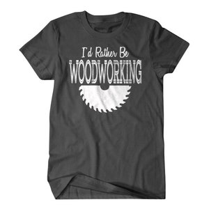 以下が含まれることがあります： 白い「I'd Rather Be Woodworking」の文字と白い鋸刃のグラフィックが描かれた濃いグレーのTシャツです。