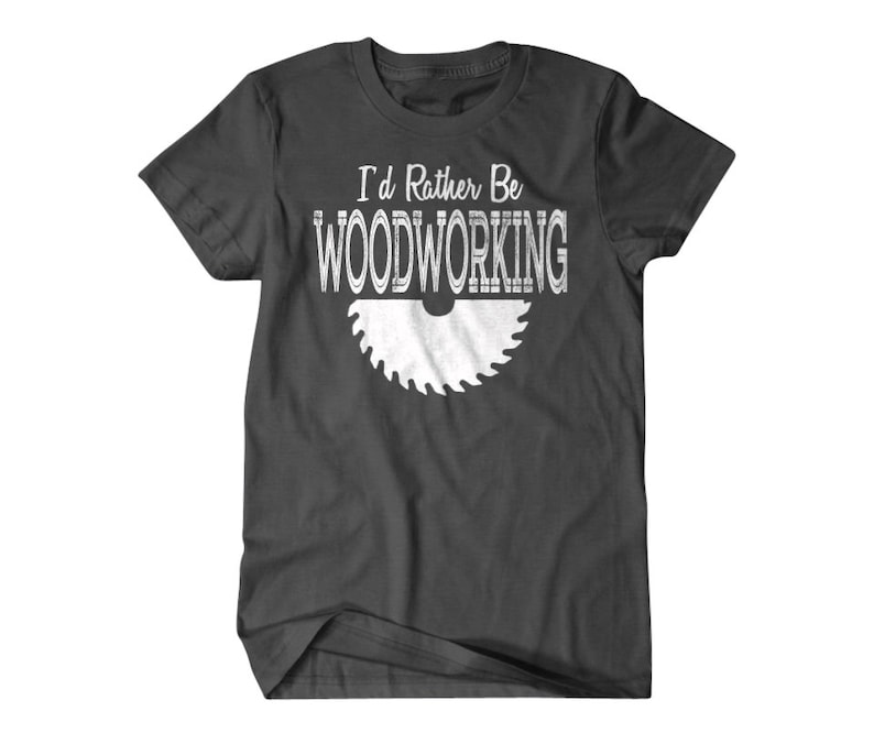 以下が含まれることがあります： 白い「I'd Rather Be Woodworking」の文字と白い鋸刃のグラフィックが描かれた濃いグレーのTシャツです。
