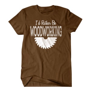 以下が含まれることがあります： 白い文字で「I'd Rather Be Woodworking」と書いてある茶色のTシャツ。白い鋸のシルエットが描かれている。