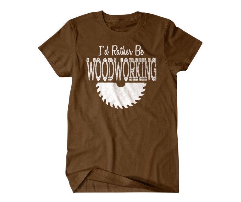 以下が含まれることがあります： 白い文字で「I'd Rather Be Woodworking」と書いてある茶色のTシャツ。白い鋸のシルエットが描かれている。