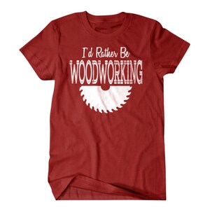 以下が含まれることがあります： 白い文字で「I'd Rather Be Woodworking」と書かれた赤いTシャツ。白いノコギリの刃のグラフィックが描かれている。