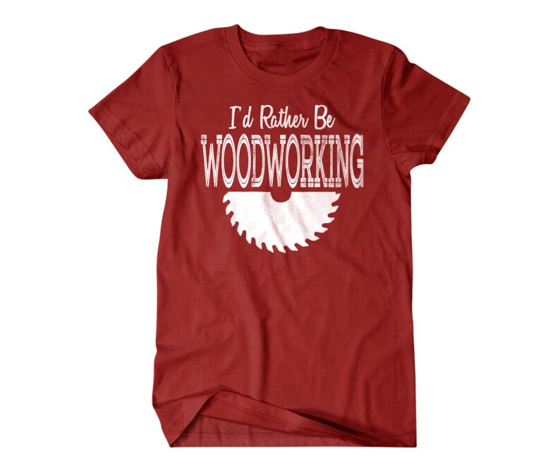 以下が含まれることがあります： 白い文字で「I'd Rather Be Woodworking」と書かれた赤いTシャツ。白いノコギリの刃のグラフィックが描かれている。