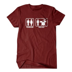 Peut inclure: Un t-shirt marron avec un graphique blanc représentant une femme en bâton en robe et le mot "PROBLÈME" en dessous. À droite, un homme en bâton avec une batterie et le mot "RÉSOLU" en dessous.