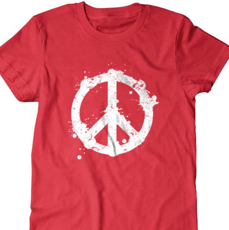 Puede incluir: Camiseta roja con un gr&aacute;fico de signo de paz blanco. El signo de paz est&aacute; rodeado de salpicaduras de pintura blanca.