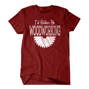 以下が含まれることがあります： 白い文字で「I'd Rather Be Woodworking」と書かれた赤いTシャツと、白い鋸刃のシルエット。
