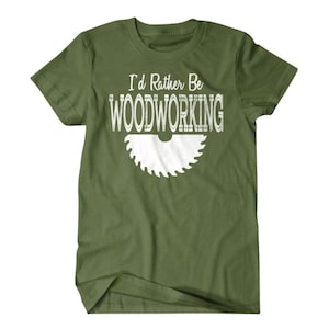 以下が含まれることがあります： 白い文字で「I'd Rather Be Woodworking」と書いてある緑色のTシャツと、白いノコギリの刃のグラフィック