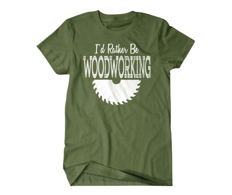 以下が含まれることがあります： 白い文字で「I'd Rather Be Woodworking」と書いてある緑色のTシャツと、白いノコギリの刃のグラフィック