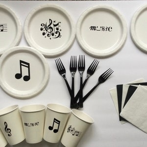 May include: A set of white paper plates and cups with black musical note designs. The plates have different designs, including a treble clef, a musical note, and the word "MUSIC". The cups have a treble clef, a musical note, and the word "MUSIC". There are also black plastic forks and white napkins.