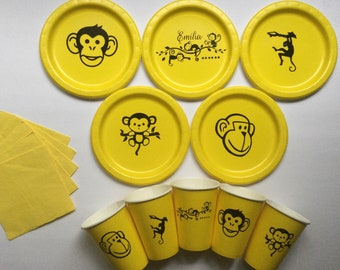 May include: Yellow paper plates and cups with black monkey designs. The plates have different monkey designs, including a monkey with a banana, a monkey hanging from a vine, and a monkey with a smile. The cups have a monkey with a banana and a monkey hanging from a vine. The text "Emilia" is on one of the plates.