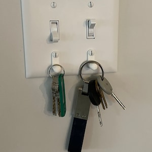 May include: White light switch plate with two toggle switches and two key hooks. A set of keys with a green key fob is hanging on one hook, and a set of keys with a blue leather key fob is hanging on the other hook.