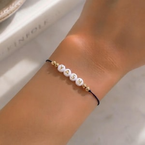 May include: A thin black cord bracelet featuring four white spherical beads with the gold-colored letters Y, A, S, and H printed on them, flanked by two small metallic gold beads on each side of the lettered sequence.