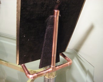 May include: A copper picture frame stand with a black velvet backing. The stand is made of copper pipe and has a simple design.