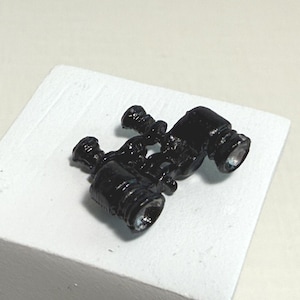 May include: A miniature black toy pair of binoculars with silver-colored lenses, resting on a white, square block. The binoculars have a detailed design, suggesting a vintage or classic style.