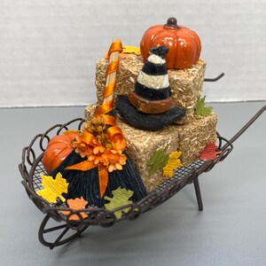 May include: A miniature Halloween-themed wheelbarrow decoration. It features a small orange pumpkin, a witch's hat, and a broom resting on hay bales. Colorful autumn leaves and flowers add to the festive fall decor.