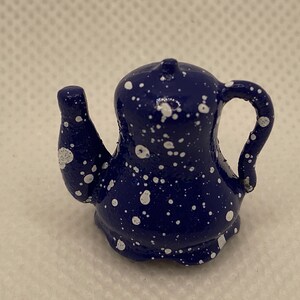 May include: A small, navy blue ceramic teapot with white speckled detailing. The teapot features a curved handle and a spout, and a small knob on the lid. The design is reminiscent of a starry night.