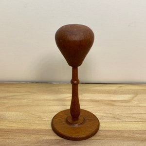May include: A wooden hat stand with a round base and a tall, slender stem. The top of the stand is shaped like a rounded dome.