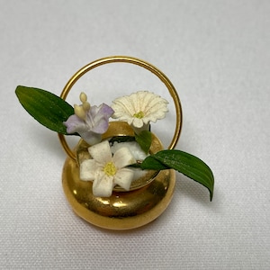 May include: A gold-colored miniature vase with a handle, filled with small white and purple flowers and green leaves. The vase has a round base and a circular handle. The flowers are detailed and delicate, creating a miniature floral arrangement.