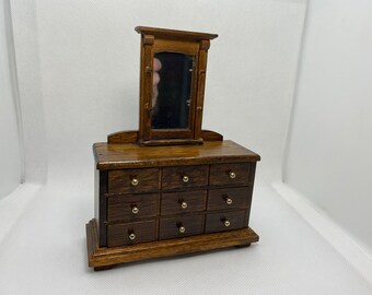 May include: Miniature wooden dresser with a mirror. The dresser has nine drawers with brass knobs and a dark brown finish. The mirror is rectangular and framed in matching wood. A charming piece for a dollhouse or display.
