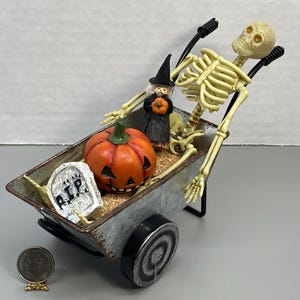 May include: A Halloween-themed miniature scene featuring a skeleton pushing a wheelbarrow filled with a carved pumpkin, a tombstone, and a witch figurine. The wheelbarrow is made of metal and has a distressed finish.
