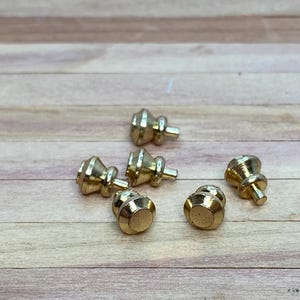 May include: Six gold-toned cabinet knobs with a rounded, slightly domed top and a small stem. The knobs are arranged on a light brown wooden surface.