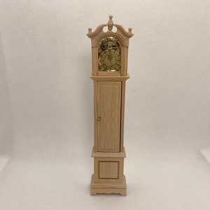 May include: A miniature wooden grandfather clock with a light finish. The clock features a gold-colored clock face and a small door. The clock is a decorative item.