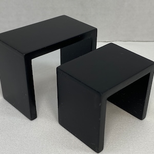 May include: Two black, U-shaped wooden display stands. The stands are rectangular and have a glossy finish. The larger stand is positioned behind the smaller one. These stands are suitable for displaying small items.
