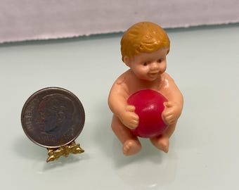 May include: A small plastic baby doll with blonde hair, holding a red ball. A U.S. dime coin is next to the doll, propped up by a gold-colored metal stand. The doll is nude and has a smiling expression.
