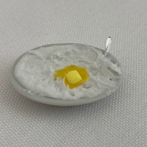 May include: Miniature bowl of mashed potatoes with a yellow pat of butter and a tiny silver spoon. The bowl is oval-shaped and has a white interior and a gray rim. The mashed potatoes are textured and fluffy.